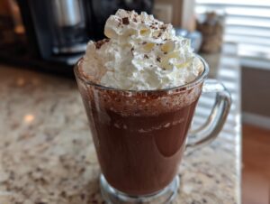 A tall glass mug filled with rich frozen hot chocolate topped high with whipped cream and chocolate shavings.