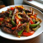 A close-up of sizzling steak fajitas featuring seasoned strips of beef mixed with vibrant red and green bell peppers and onions.
