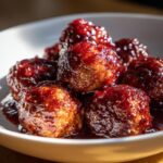 A close-up of several glazed cocktail meatballs piled in a white bowl, covered in a thick, dark red sauce.