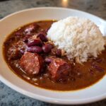 A close-up of a white bowl filled with rich, savory red beans and rice, featuring chunks of smoked sausage.