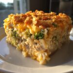 A square serving of creamy rice casserole with broccoli, cheese, and a crunchy topping on a white plate.
