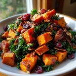 A vibrant bowl of sweet potato salad featuring roasted orange sweet potato cubes, dark green kale, pecans, and dried cranberries.