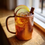 A frosty copper mug filled with an apple cider Moscow mule, garnished with apple slices and a cinnamon stick.