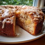A close-up of a slice removed from a homemade apple crumb cake topped with cinnamon streusel and vanilla glaze.