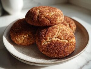 A close-up of four freshly baked Snickerdoodle cookies, richly coated in cinnamon sugar, stacked on a speckled ceramic plate.