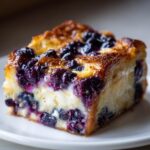 A close-up of a rich, golden-brown slice of Blueberry French Toast Casserole with creamy filling and burst blueberries.