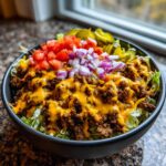 A close-up of a fully loaded cheeseburger bowl with seasoned ground beef, melted cheese, lettuce, tomatoes, onions, and pickles.