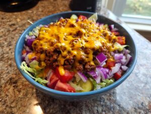 A vibrant cheeseburger bowl featuring seasoned ground beef, melted cheddar cheese, lettuce, tomatoes, and red onions.