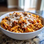 A close-up of a white bowl filled with cheesy enchilada pasta featuring rotini noodles and a meat sauce.