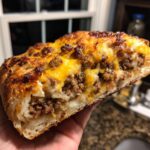 A hand holding a thick slice of baked garbage bread stuffed with seasoned ground meat and melted cheddar cheese.