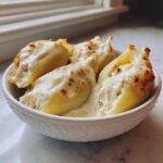 Four baked chicken alfredo stuffed shells covered in melted, slightly browned cheese sauce in a white bowl.