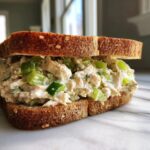 A close-up of a generously filled chicken salad sandwich on toasted whole wheat bread, showing creamy filling with green celery chunks.
