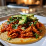 A single corn tortilla topped with saucy, shredded chicken tinga, fresh cilantro, and sliced avocado.