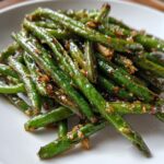 A close-up of vibrant green, blistered chinese green beans with garlic sauce piled on a white plate.