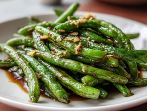 A close-up of bright green, glossy chinese green beans with garlic sauce piled on a white plate.