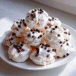 A pile of fluffy, white meringue forgotten cookies topped with mini chocolate chips served on a white plate.