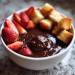Close-up of a white bowl filled with rich chocolate fondue, surrounded by fresh sliced strawberries and golden cake cubes.