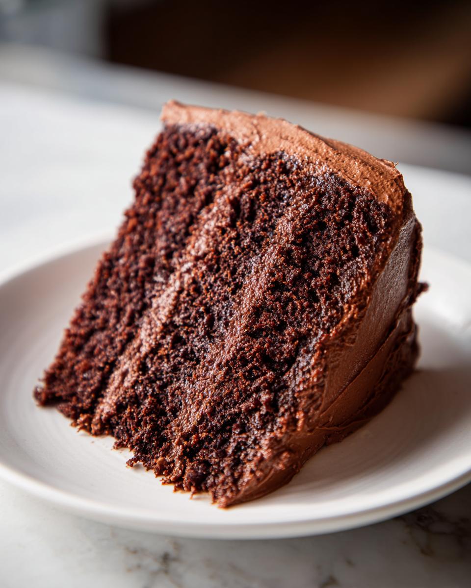 A close-up of a moist, dark chocolate slice of capricorn cake with thick chocolate frosting on a white plate.