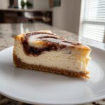 A perfect slice of cinnamon roll cheesecake featuring a creamy center, cinnamon swirl, and white icing on a white plate.