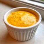 Close-up of a small white ramekin filled with bright orange, creamy cottage cheese queso dip speckled with spices.