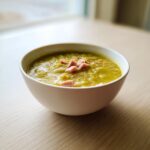 A close-up of a white bowl filled with thick, green pea soup topped with diced ham.