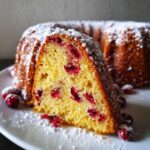 Close-up of a slice of moist cranberry orange cake, studded with red cranberries and dusted heavily with powdered sugar.