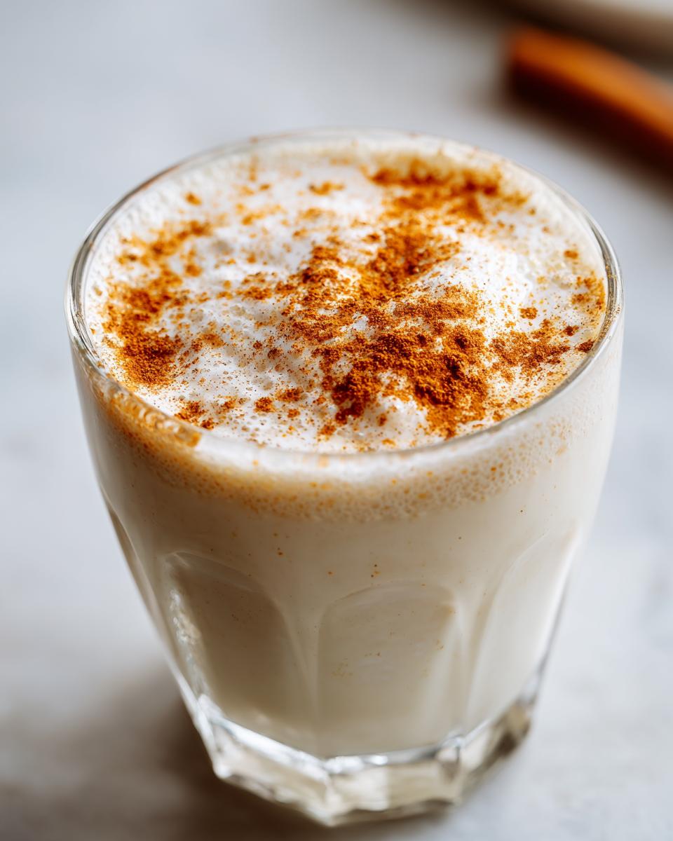Close-up of a glass filled with creamy coquito, topped with frothy foam and sprinkled with cinnamon.