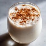 Close-up of a glass filled with creamy, frothy coquito, generously dusted with ground cinnamon on top.