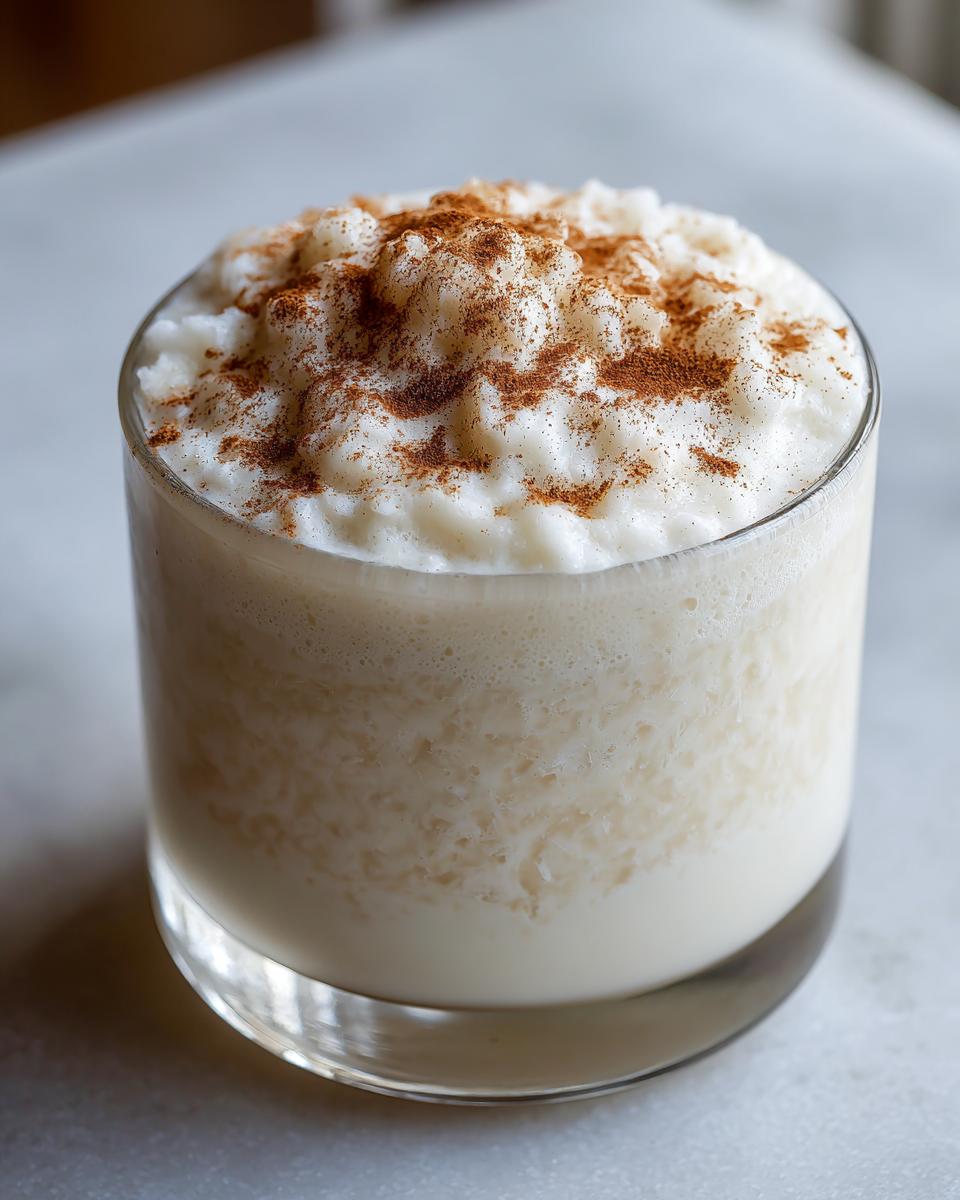 Close-up of a glass of creamy coquito topped with rice pudding texture and dusted with ground cinnamon.
