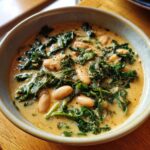 A close-up bowl of creamy tuscan white bean soup featuring large white beans and wilted kale.