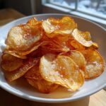 A close-up of a white bowl filled with golden brown, crispy homemade potato chips sprinkled with coarse sea salt.