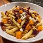 A close-up of a bowl filled with fall pasta salad featuring penne, roasted butternut squash, pecans, dried cranberries, and feta cheese.