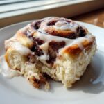 A close-up shot of a single, fluffy cinnamon roll topped with thick white icing, sitting on a white plate.