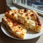 Close-up of fluffy homemade naan bread stacked on a white plate, showing airy texture and golden-brown charred spots.