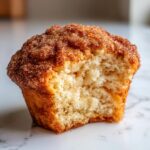 A close-up of a delicious snickerdoodle muffin with a bite taken out, showing the soft interior and cinnamon sugar topping.