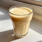 A tall glass filled with a creamy, light orange banana smoothie, sitting on a white surface in bright sunlight.
