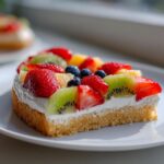 A close-up slice of fruit pizza sugar cookie topped with cream cheese frosting, strawberries, kiwi, and blueberries.