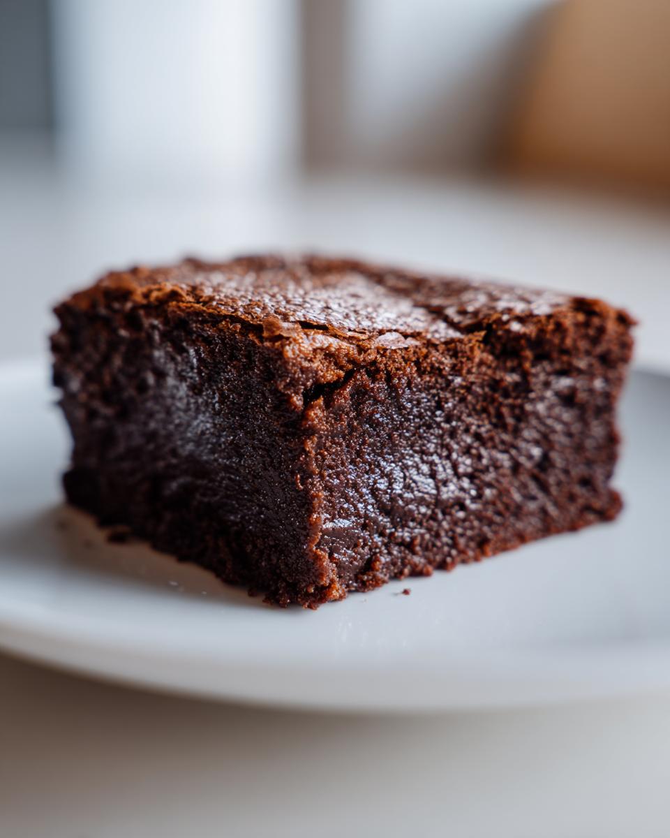 A close-up, macro shot of a perfectly baked, rich, dark chocolate fudgy brownie sitting on a white plate.