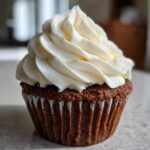 A close-up of a rich, dark gingerbread cupcakes topped with a tall swirl of white cream cheese frosting.
