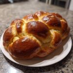 A beautifully braided, golden-brown, shiny 3-strand Challah loaf sprinkled with sesame seeds, resting on a white plate.