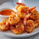 A pile of golden brown, crispy coconut shrimp served on a white plate next to a small bowl of sweet chili dipping sauce.