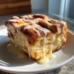 A decadent, square serving of Cinnamon roll casserole topped with thick vanilla icing dripping down the sides.