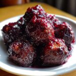 A close-up of savory meatballs coated thickly in a glossy, dark purple grape jelly sauce, ready to serve.