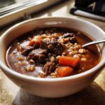 Close-up of a rich, dark bowl of beef barley soup featuring tender beef chunks, plump barley, and bright orange carrots.