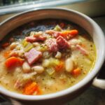 Close-up of a bowl of thick navy bean soup featuring chunks of ham, carrots, and celery.