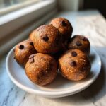 A stack of homemade, textured protein balls topped with mini chocolate chips, resting on a white plate near a window.