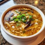 A close-up of a steaming bowl of hot and sour soup recipe, garnished with fresh green onions and mushrooms.