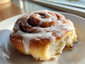 A close-up, sunlit photo of a fluffy, freshly baked cinnamon roll generously topped with white icing.