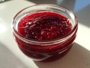 Close-up view of a small glass jar filled with glistening, deep red christmas jam, showing visible seeds.