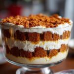 Close-up of a beautiful gingerbread trifle featuring layers of brown cake cubes, white cream, and a crumb topping.